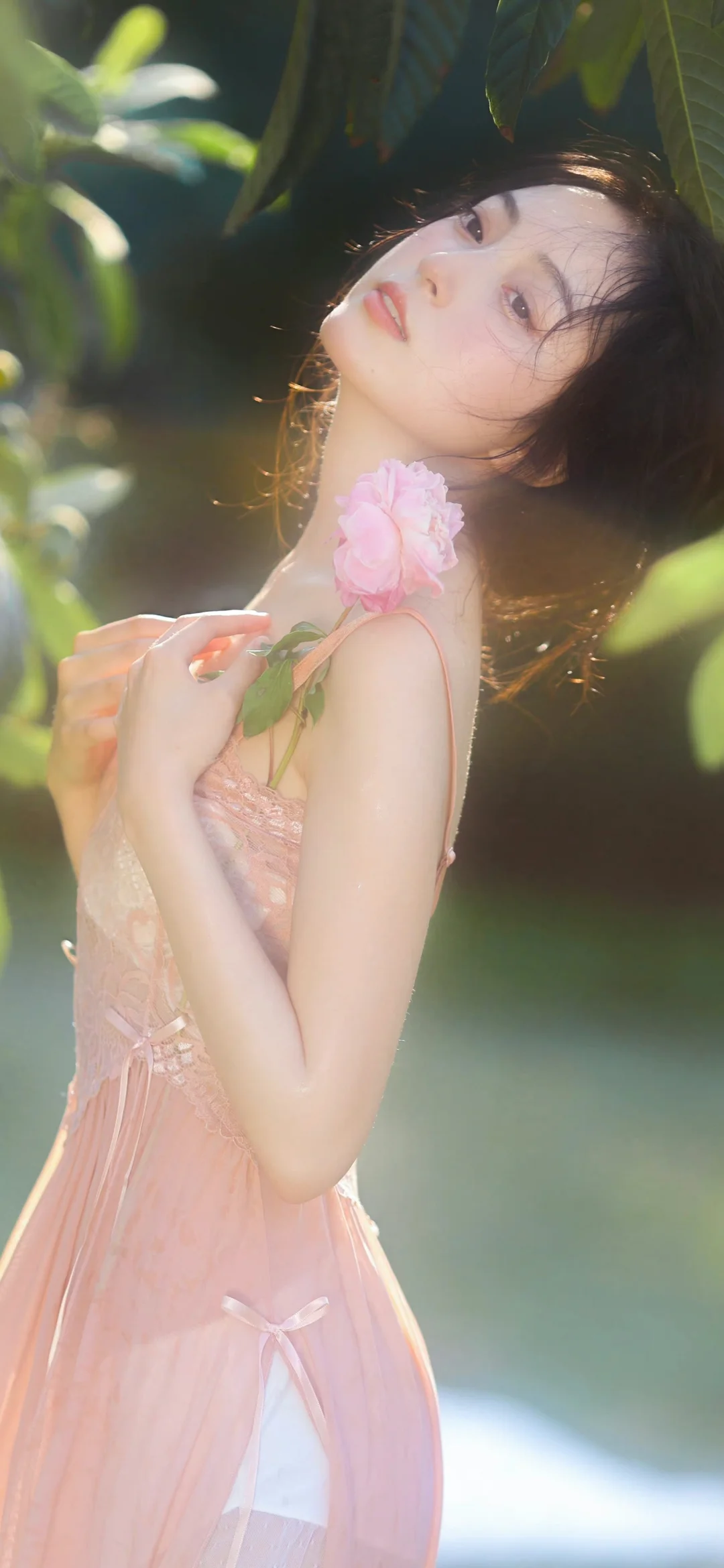 一名身穿粉色连衣裙的女子手持粉色花朵，站在阳光映照的绿叶间。
