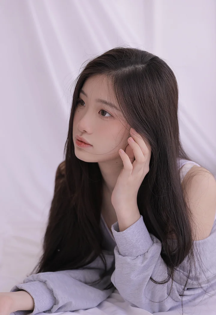 긴 머리의 여성이 부드러운 연보라색 배경 앞에서 옆을 바라보고 있다.