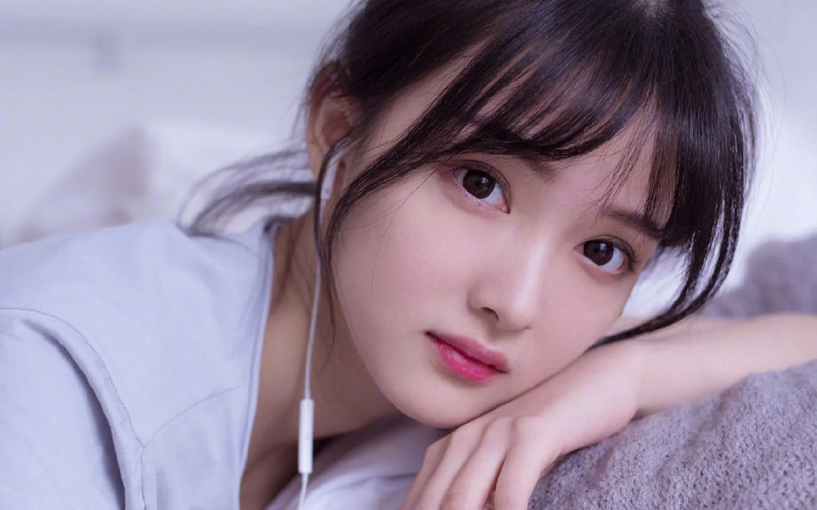 一名女子穿着白色上衣，戴着有线耳机，靠在柔软表面上看向镜头。