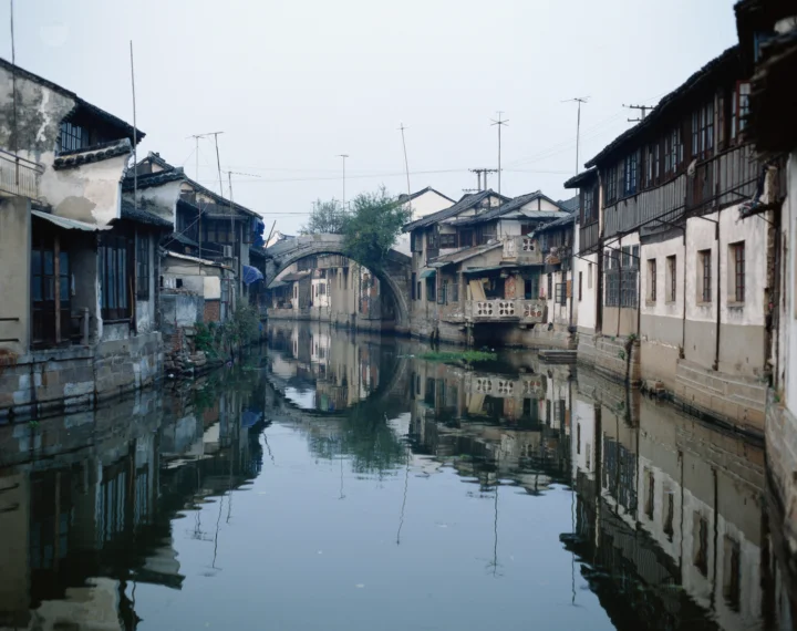 一条宁静的水道穿过两岸相连的传统民居，远处可见一座石桥，建筑倒映在水面上。