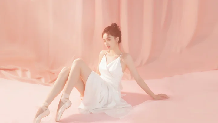 흰색 드레스를 입은 성인 여성이 분홍색 배경 앞에 앉아 있습니다.