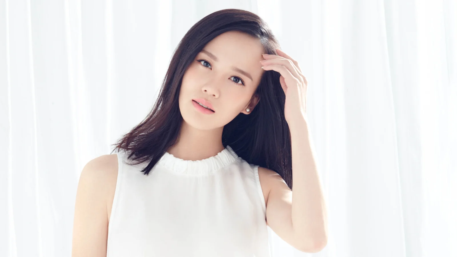 흰색 상의를 입은 성인 여성이 밝은 흰색 커튼 앞에서 머리카락을 만지고 있다.