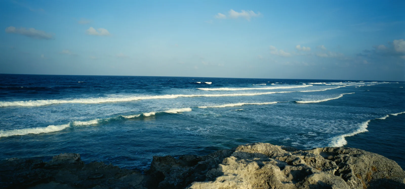 蓝色海面上的层层浪涛拍打着前景的岩石海岸，天空晴朗开阔。