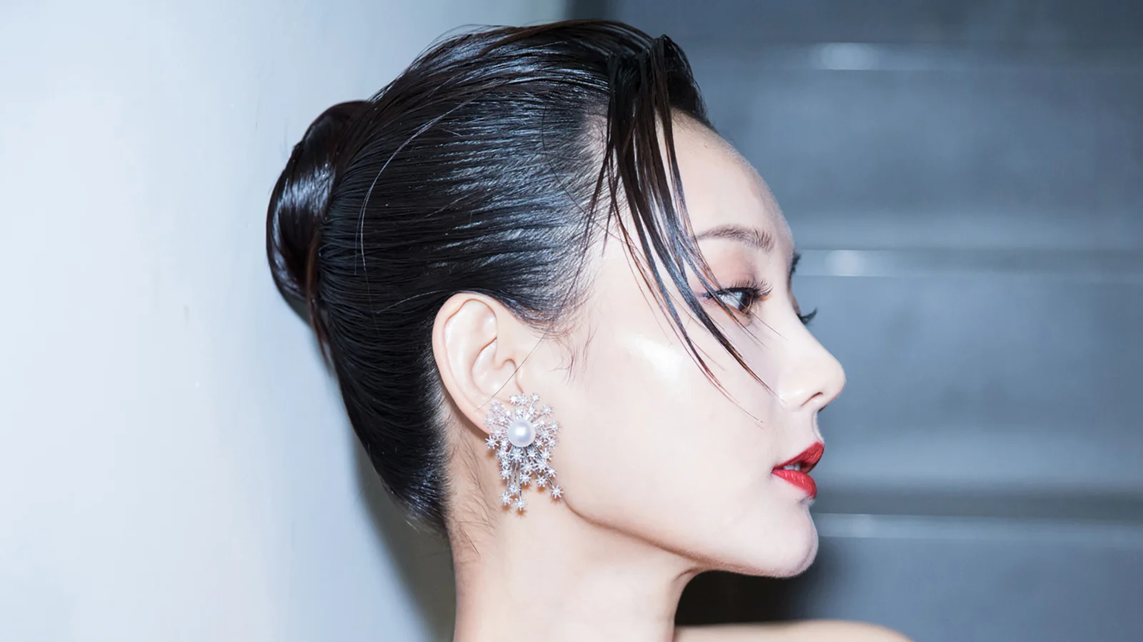 一位扎着深色发髻、佩戴华丽耳饰的成年女性侧脸出现在冷蓝灰色背景前。