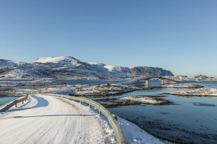 积雪覆盖的山脉环绕着峡湾水面，一条蜿蜒道路通向远处的桥梁，天空湛蓝。