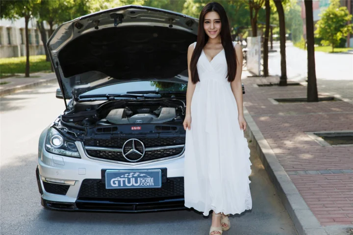 一名身穿白色长裙的成年女子站在一辆打开引擎盖的银色汽车旁，背景是阳光下的林荫街道。