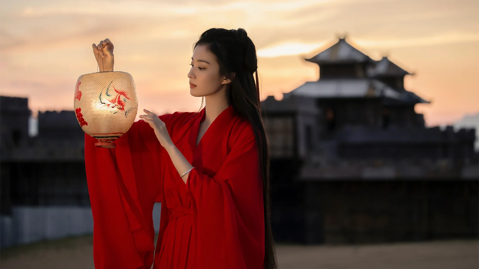 一名身穿红色长袍的女子在夕阳下手持绘有图案的灯笼，背景是传统建筑。