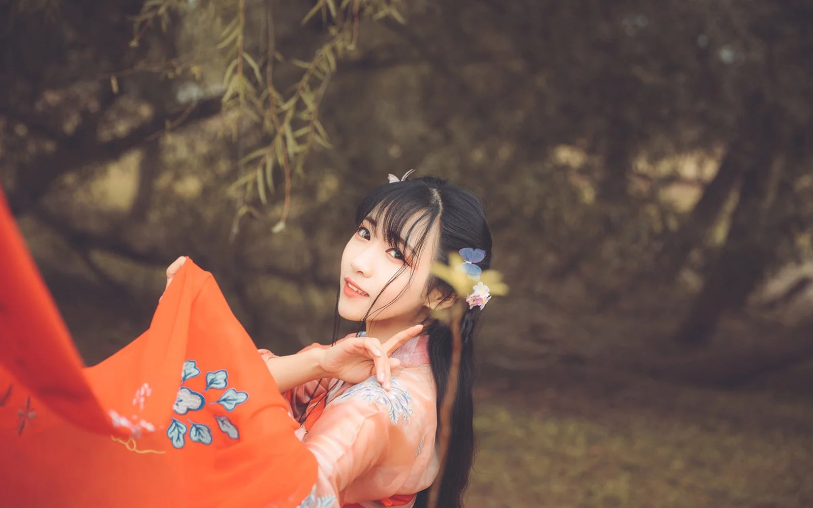 주황색 전통 의상을 입은 여성이 나무를 배경으로 옆을 향해 미소 짓고 있다.
