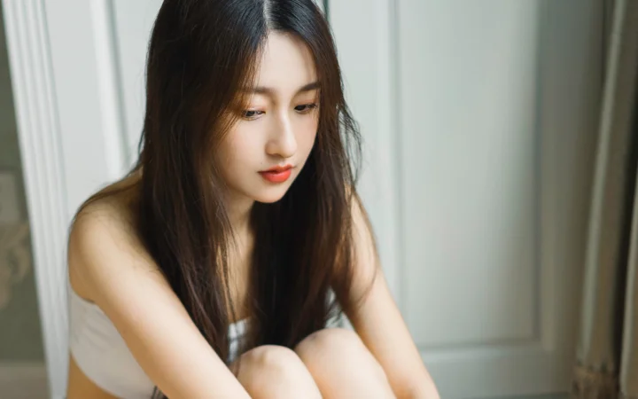 一名长发女性坐在室内窗边，低头看向一侧。