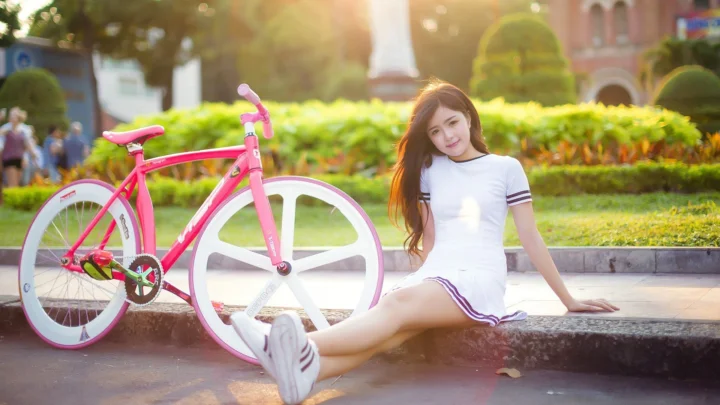 一名年轻女子坐在公园路边，身旁停着一辆粉色自行车，阳光从后方洒落。