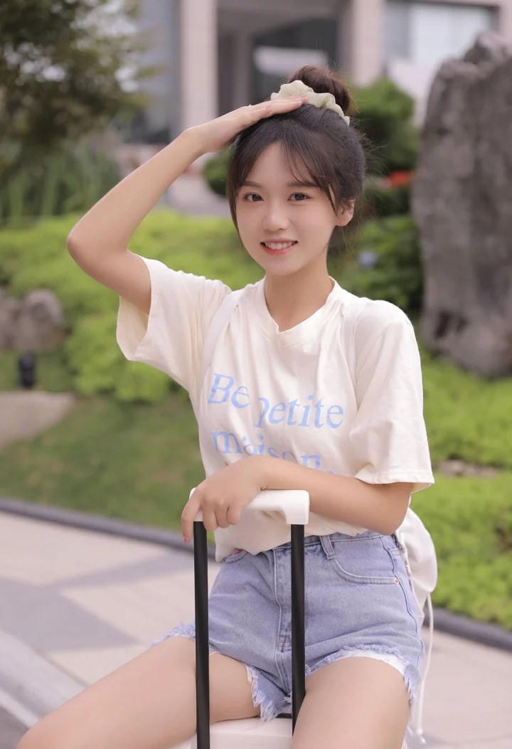一名身穿白色T恤和牛仔短裤的女性在户外手扶行李箱并整理头发。