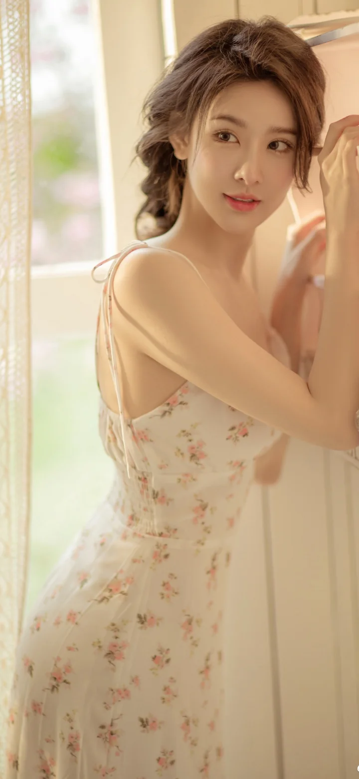 一名成年女性穿着碎花连衣裙站在明亮窗边回望镜头。