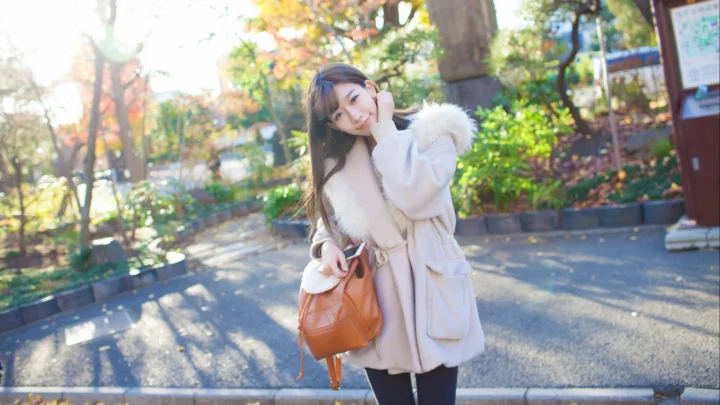 밝은 색 코트를 입은 성인 여성이 갈색 핸드백을 들고 햇살이 비치는 공원 산책로 옆에서 미소 짓고 있습니다.