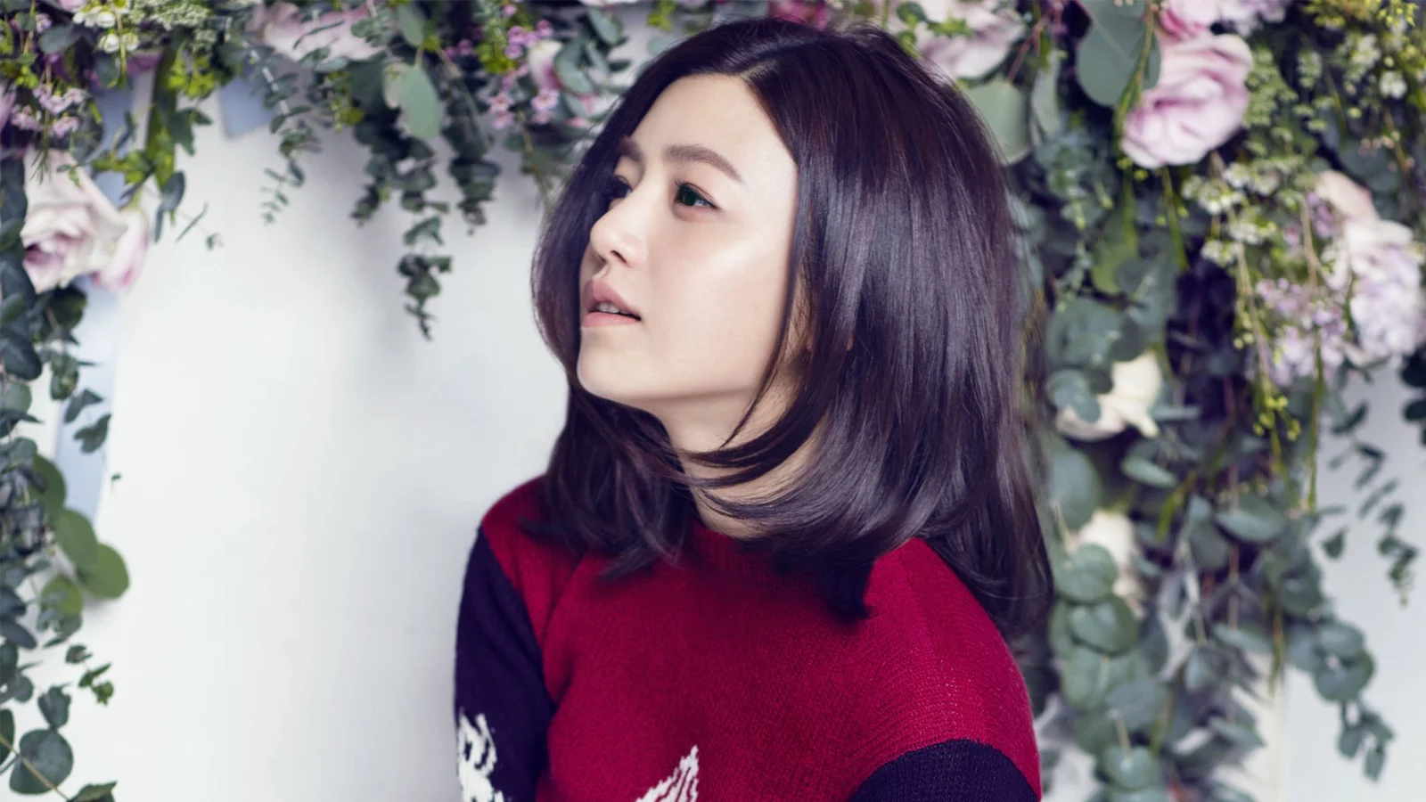 꽃과 초록 잎으로 장식된 벽을 배경으로 성인 여성이 옆모습을 보이고 있다.