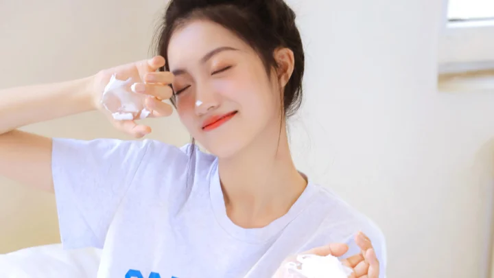 연보라색 티셔츠를 입은 여성이 눈을 감고 미소 지으며 실내에서 두 손을 얼굴 가까이에 대고 있습니다.