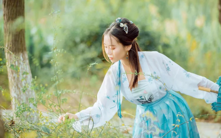 一名身穿浅色传统服饰的女子在绿意盎然的林间俯身触碰植物。
