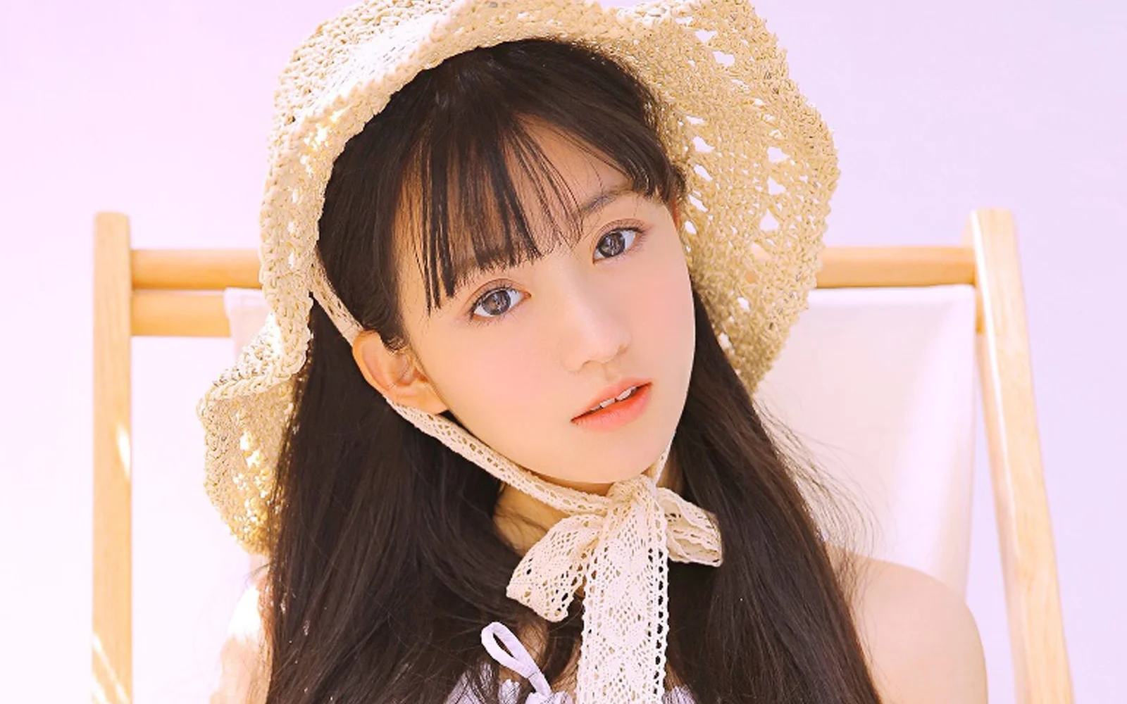 麦わら帽子とレースのリボンを身につけた女性が、淡いピンク色の背景でカメラに近づいて写っています。