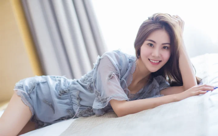 一名成年女性穿着浅蓝色连衣裙，微笑着侧躺在窗边的床上。