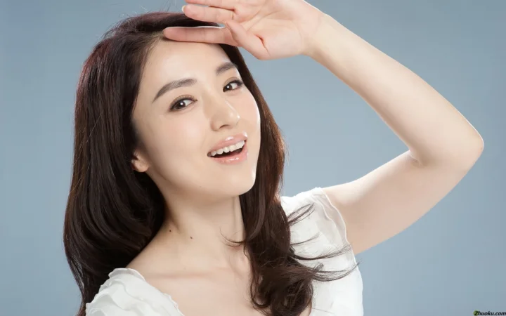 一位成年女性在浅灰色背景前微笑，并抬手遮挡眼睛上方的光线。