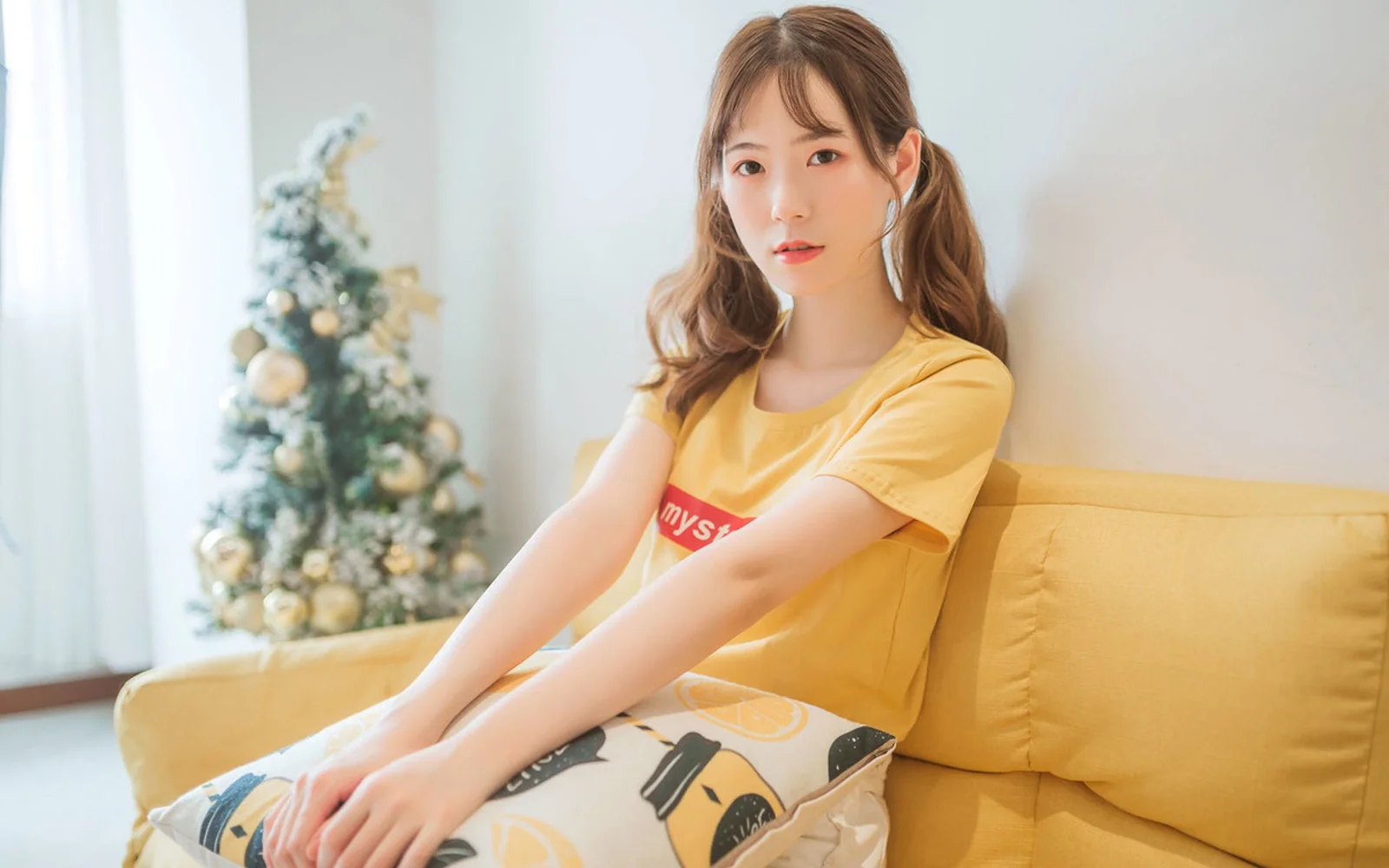 一名身穿黄色上衣的女性坐在室内黄色沙发上，背景可见一棵装饰树。
