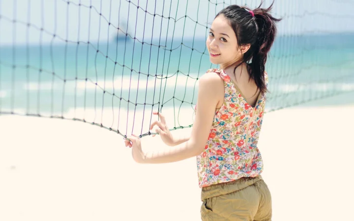 一名身穿花色上衣和休闲短裤的成年女性站在海边排球网旁回头微笑。