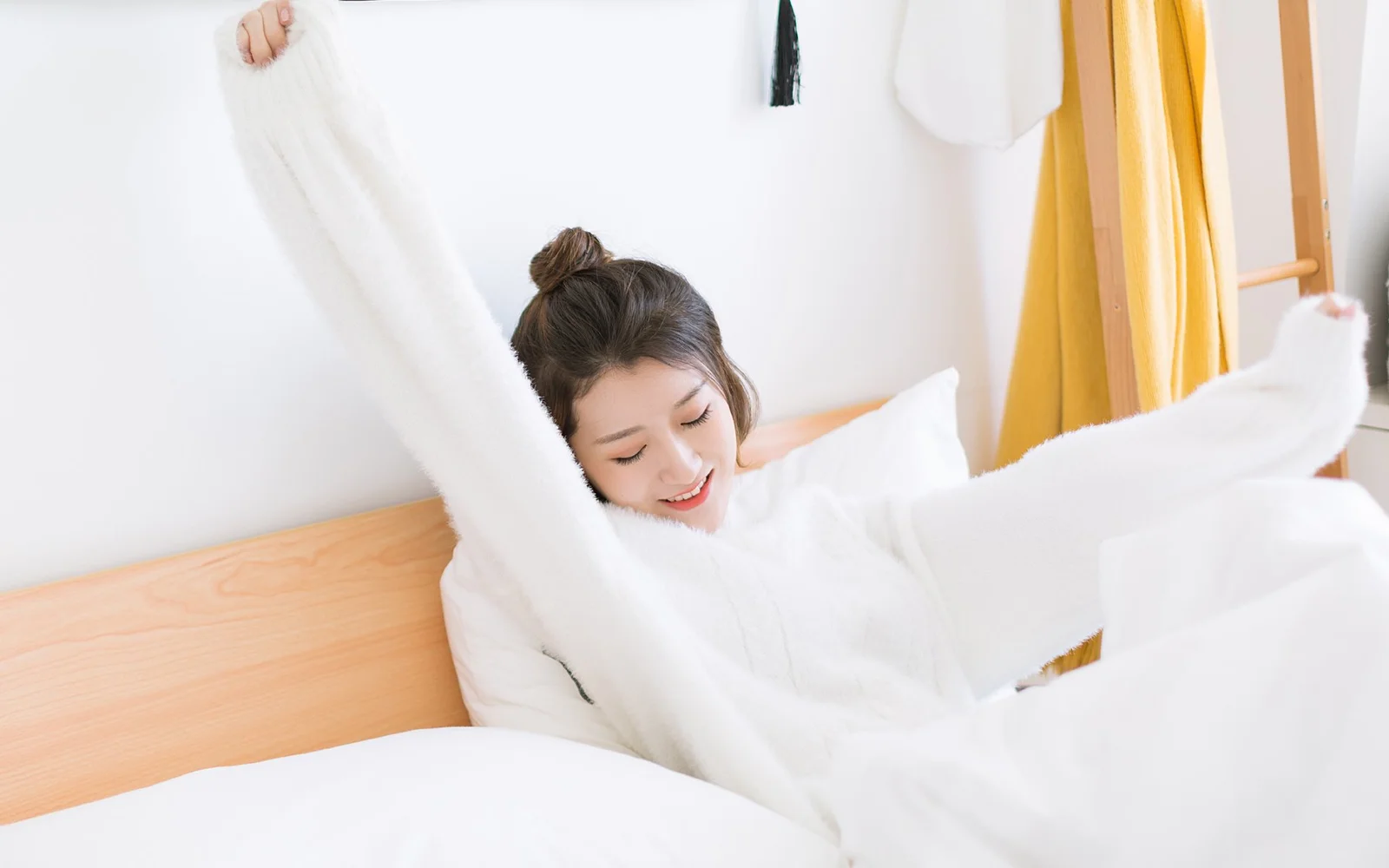 一名穿白色上衣的女性在明亮卧室的床上伸展双臂。