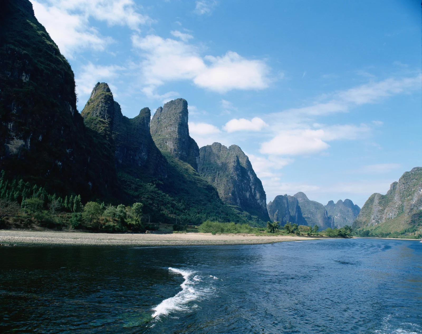蓝天下的群山峡谷环抱着宽阔河流，前景可见船尾划过的水痕。