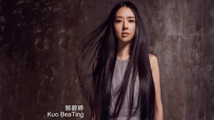 一名长发女性身穿灰色无袖上衣站在深色纹理背景前。