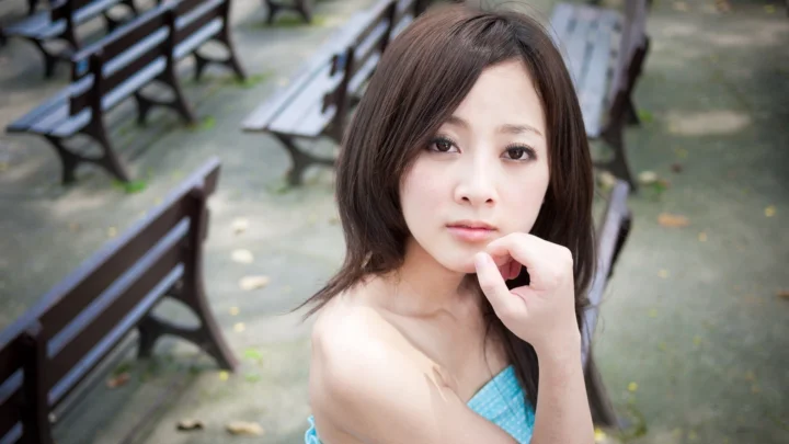 一名成年女性在户外长椅旁看向镜头，手指轻触下巴。