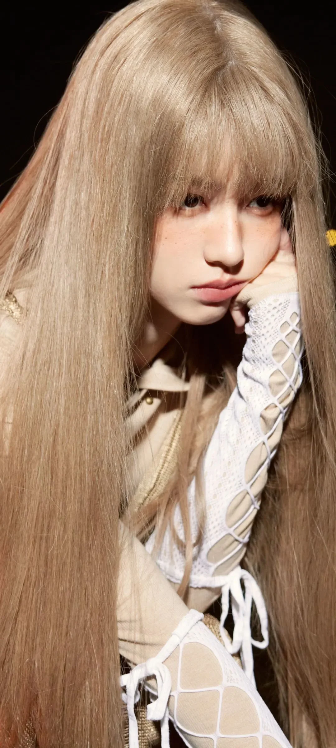 一名长发女性以手托脸，身穿浅色时尚服装，在黑色背景前拍摄。