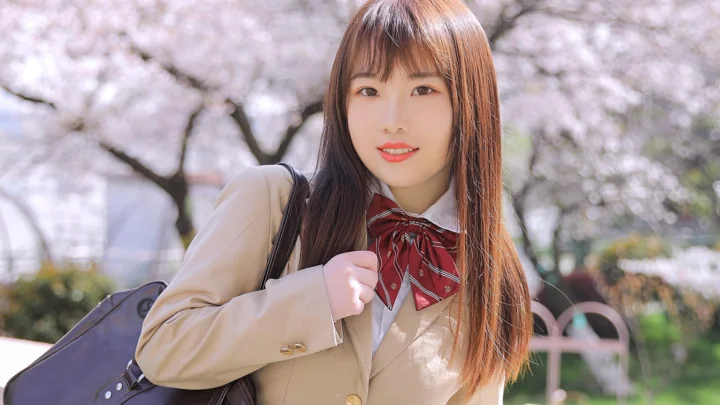 一名长发女子站在盛开的樱花树下，身穿浅色外套并背着包。