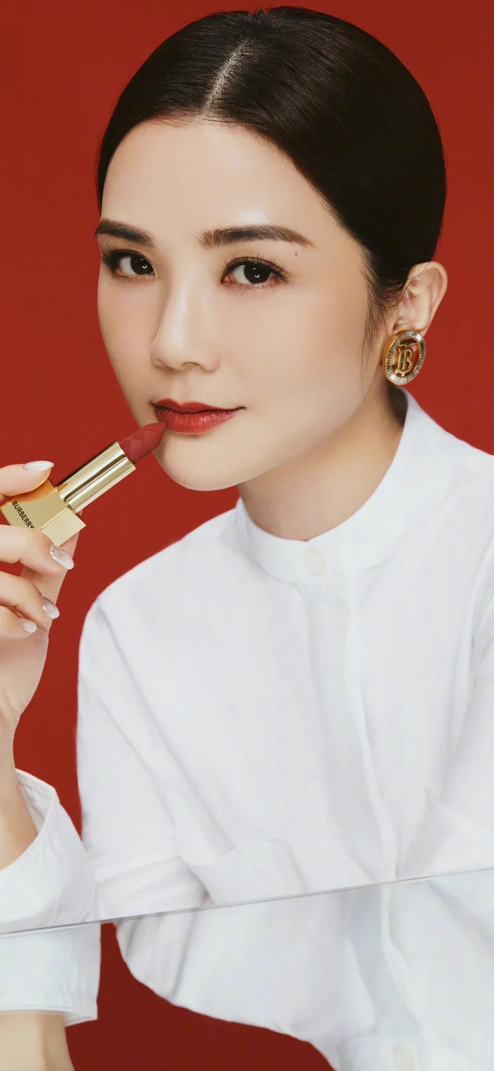 一位穿白色上衣的女性在红色背景前手持口红凝视镜头。