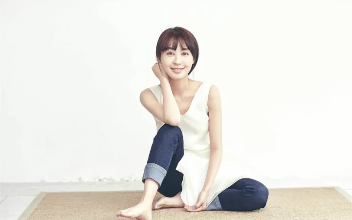흰색 민소매 상의를 입은 성인 여성이 밝은 흰색 벽을 배경으로 연한 색 러그 위에 앉아 있습니다.