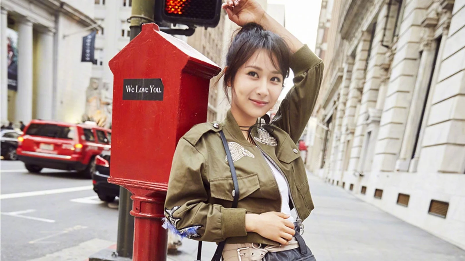一名成年女子站在城市街頭的紅色郵筒旁，背景可見建築、車輛和人行道。