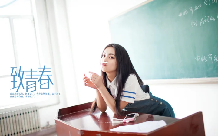 一名女性伏在教室课桌上，身后是黑板和明亮的窗户。
