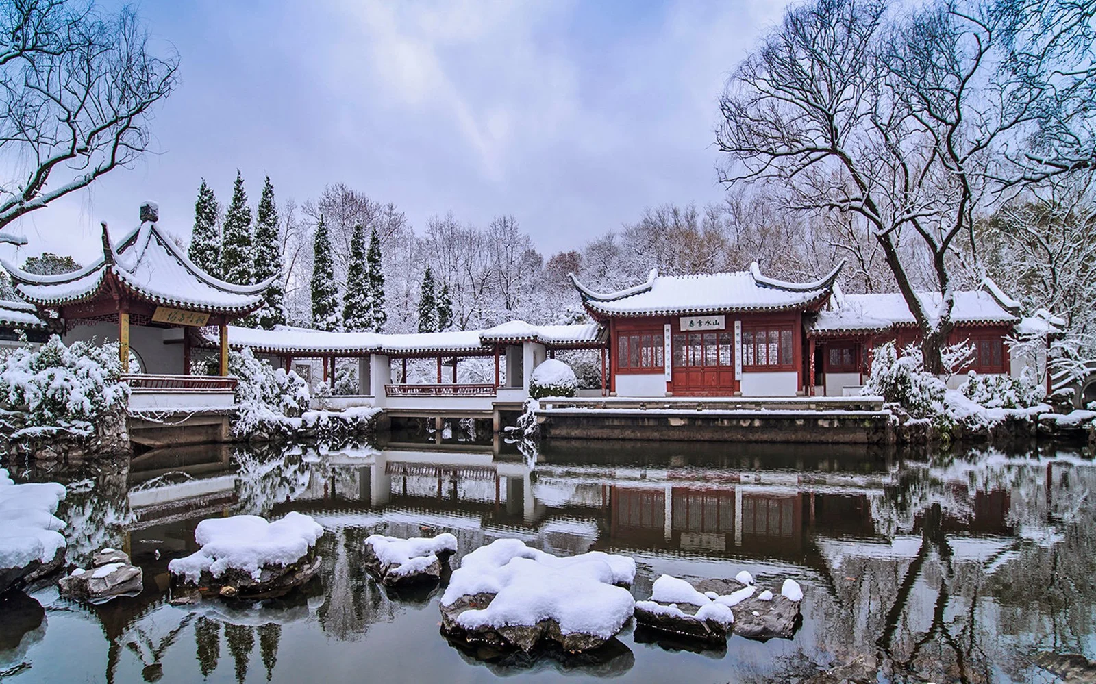 雪に覆われた中国庭園の建物が池のほとりに佇み、静かな水面に冬の景色が映っています。