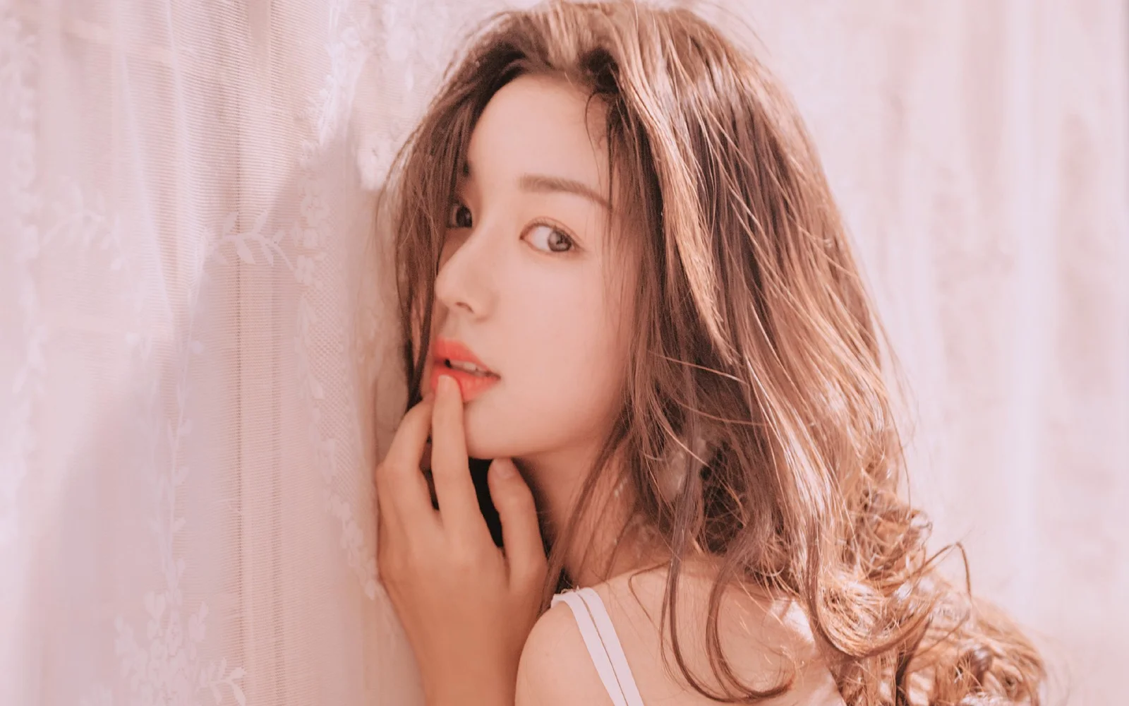 긴 머리의 성인 여성이 부드러운 분홍빛 조명 속에서 옆모습으로 카메라를 바라보고 있습니다.