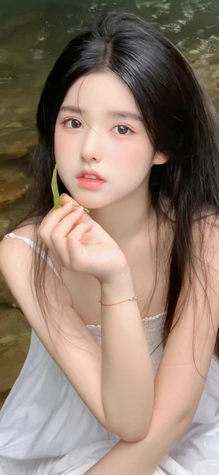 긴 머리의 여성이 개울가에서 손으로 턱을 괴고 카메라를 바라보고 있다.