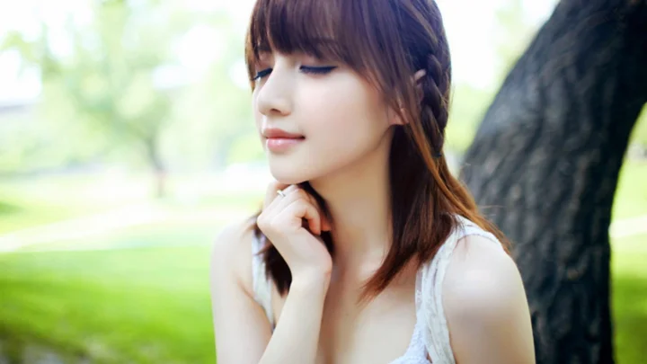 一名成年女子在绿地旁的树边闭目而立，阳光柔和，画面背景呈现清新的户外景色。