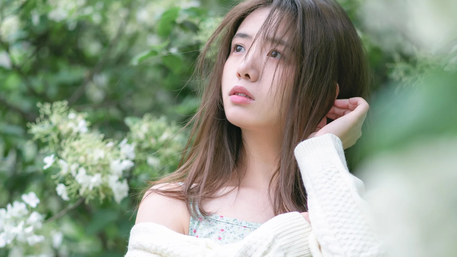 푸른 식물이 우거진 야외에서 갈색 긴 머리의 여성이 머리를 살짝 만지고 있다.