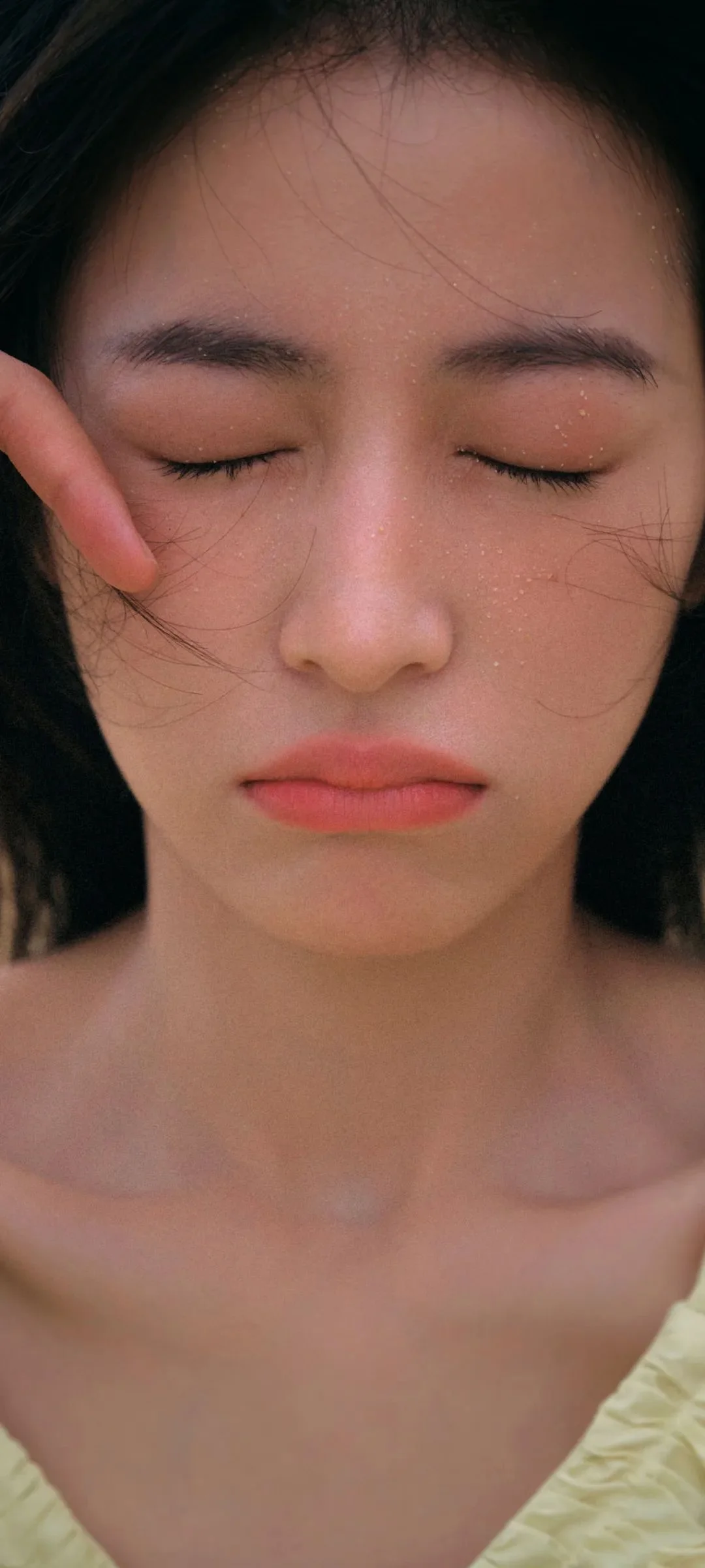目を閉じた女性の顔を近距離から捉えたポートレートで、頬のそばに手があり、画面下部に肩が見える。