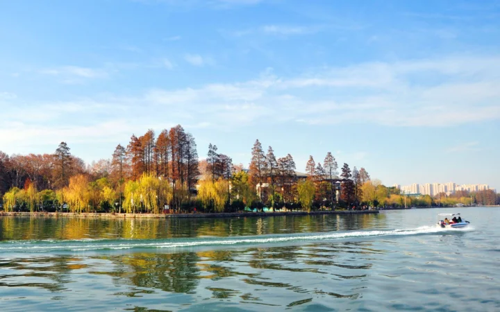 晴朗天空下，湖面映照着秋日树木与岸边景色，远处可见一艘小艇和城市建筑。