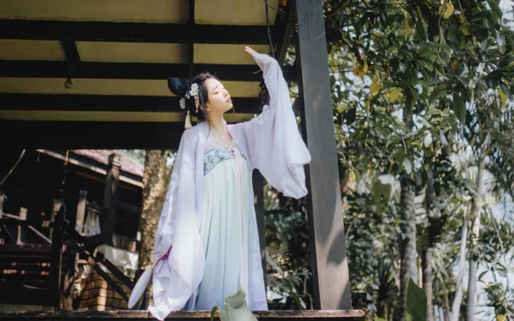 밝은 색상의 전통 의상을 입은 여성이 나무로 된 구조물 옆에 나무들로 둘러싸여 서 있습니다.