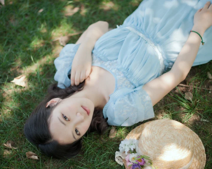 一名身穿浅蓝色连衣裙的女子躺在阳光斑驳的草地上。