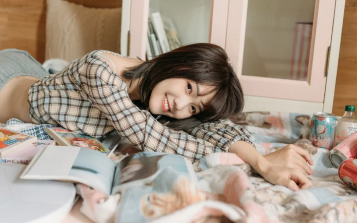一名微笑的女性穿着格纹衬衫趴在床上，身旁放着一本打开的杂志。