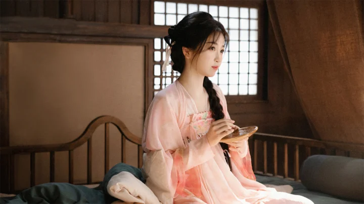 一名身穿浅色传统服饰的女子坐在木质室内空间中，手持碗具面向一侧。