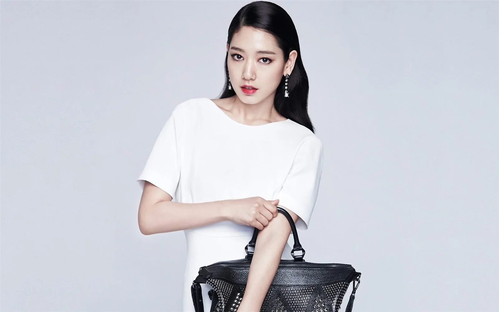 흰색 상의를 입은 여성이 연한 회색 배경 앞에서 검은색 핸드백을 들고 있다.