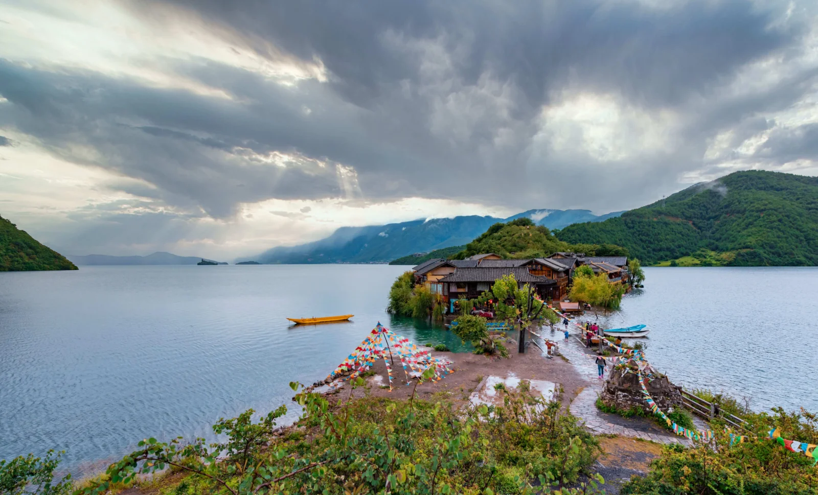 湖水延伸至远处群山之间，岸边坐落着木质建筑，厚重云层覆盖天空。