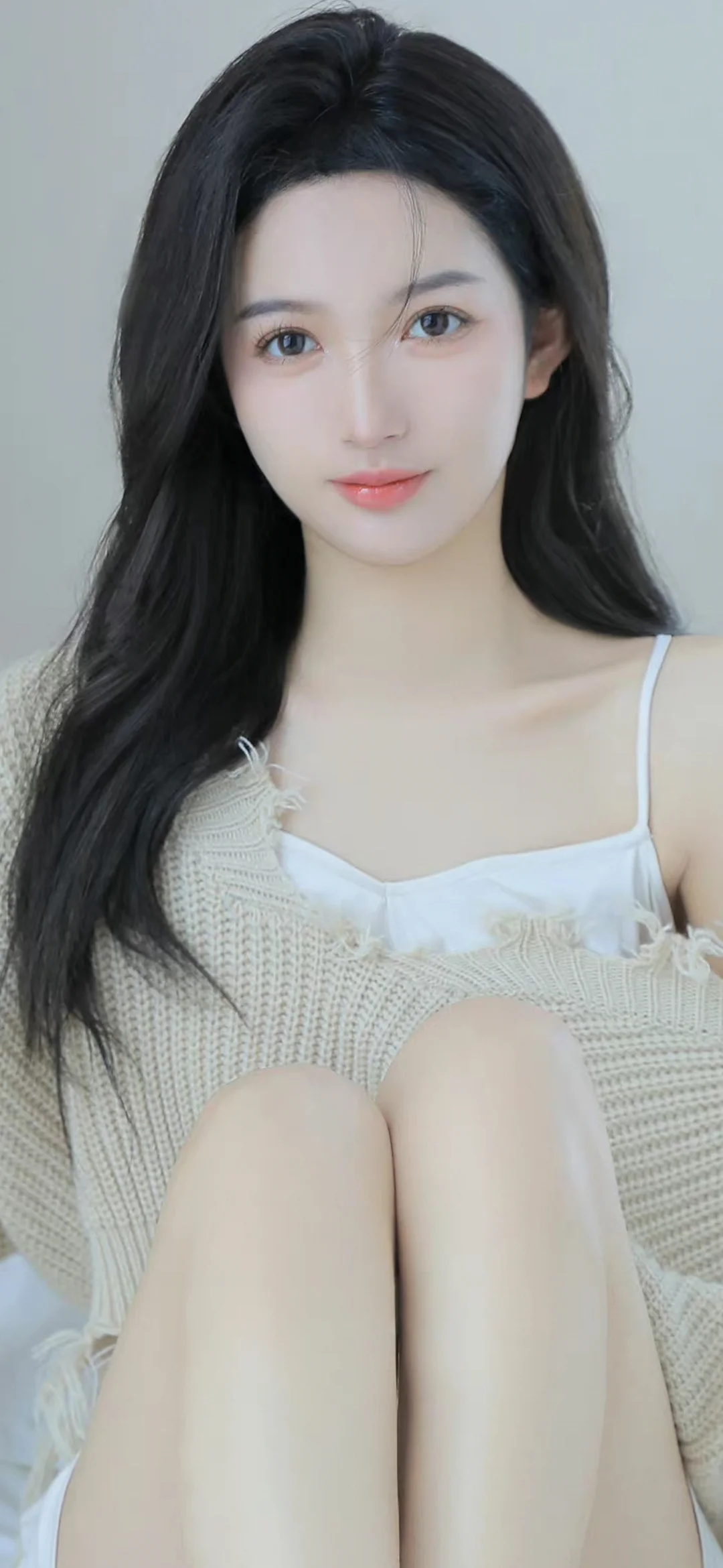 一名长发女性身穿浅色针织衫和白色吊带上衣，面向镜头坐在浅灰色背景前。