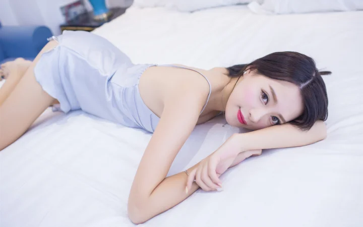 一名成年女子穿着浅色无袖上衣，侧躺在白色床上看向镜头。